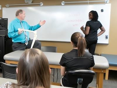 教授拿着骷髅腿模型，在课堂前和一个在白板上写字的学生交谈